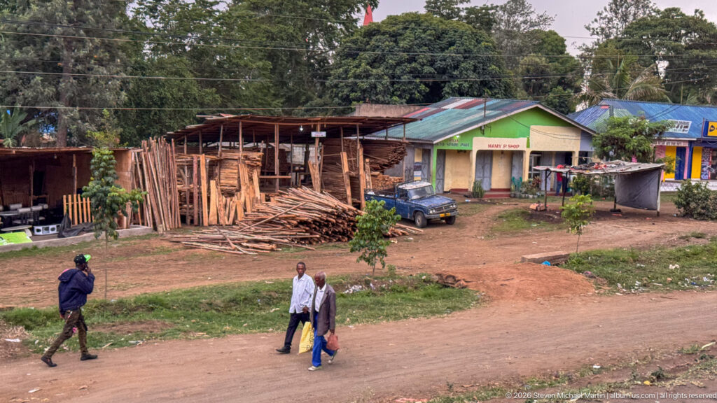 Roadside timber yard stacked with rough poles and lumber, with three pedestrians walking along the dirt verge.