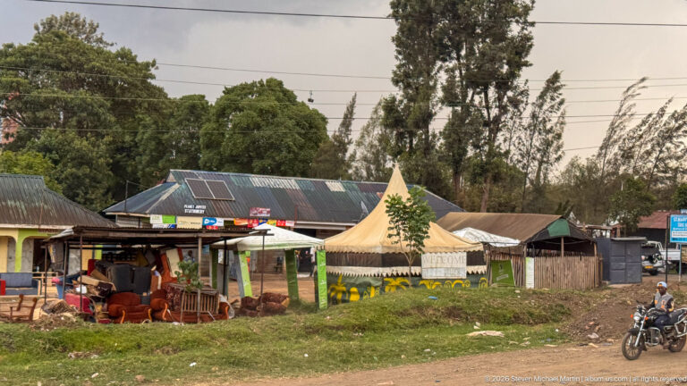 Small roadside café with open seating, a peaked canvas shelter, corrugated roof and solar panel, with a motorcyclist nearby.