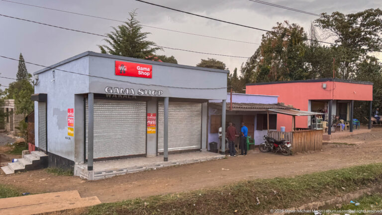 Gray Game Shop Wakala storefront with closed roller shutters beside a red roadside business, motorcycles, and several people.