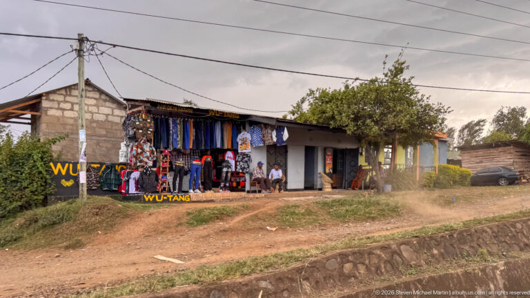 Open roadside clothing shop displaying hanging garments and Wu-Tang branding, with several people seated near the entrance.