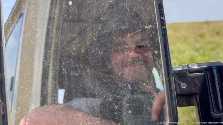 Passenger visible through a dusty safari-vehicle window, with one hand raised near the glass and grassland beyond.