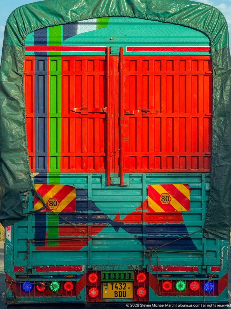Rear of a brightly painted freight truck with red doors, turquoise body, green tarp and Tanzanian license plate.
