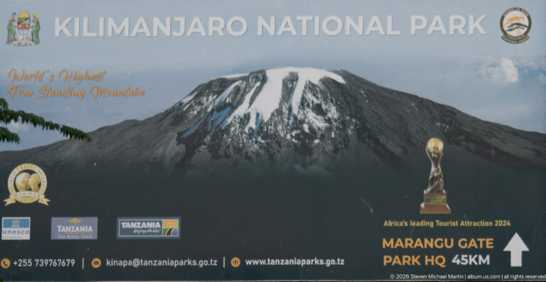 Close view of Kilimanjaro National Park sign showing mountain image, logos and Marangu Gate Park HQ 45 km arrow.