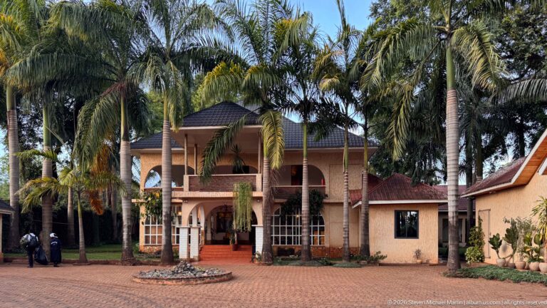 Centered view of a two-story guesthouse façade framed by palms, with arched openings, red steps and paved forecourt.