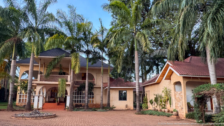 Palm-lined forecourt before a two-story cream guesthouse with arched openings, dark roof and red entrance steps.