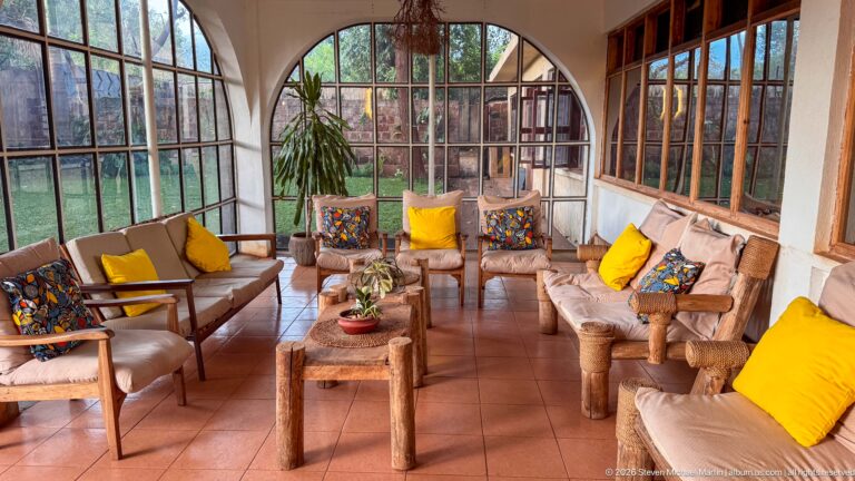 Guest lounge with rustic wooden seating, yellow cushions, terracotta floor and large arched garden windows.