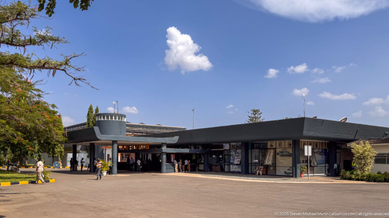 Low terminal entrance at Kilimanjaro International Airport with broad canopy, paved forecourt and people near the doors.