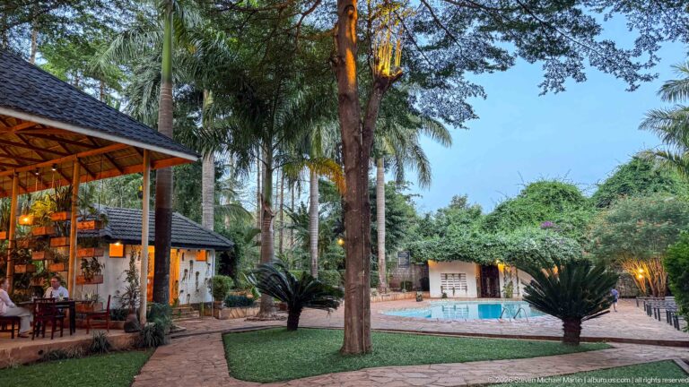 Garden path between an open dining pavilion and small swimming pool, surrounded by palms, trees and cycads.