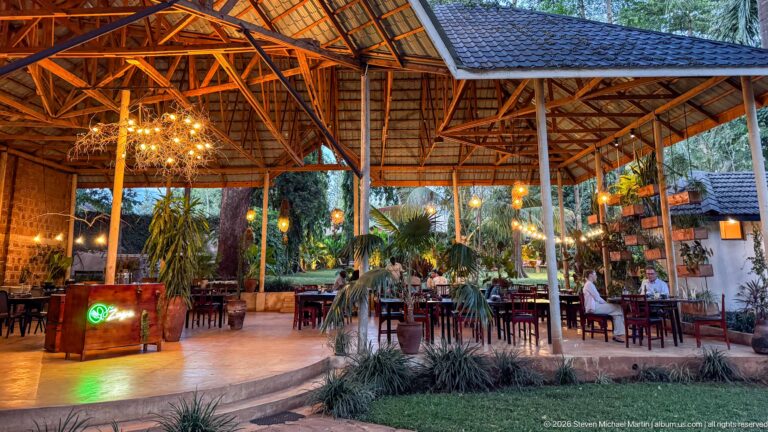 Open-air dining pavilion with exposed timber roof, hanging lights, tables and tropical plants.