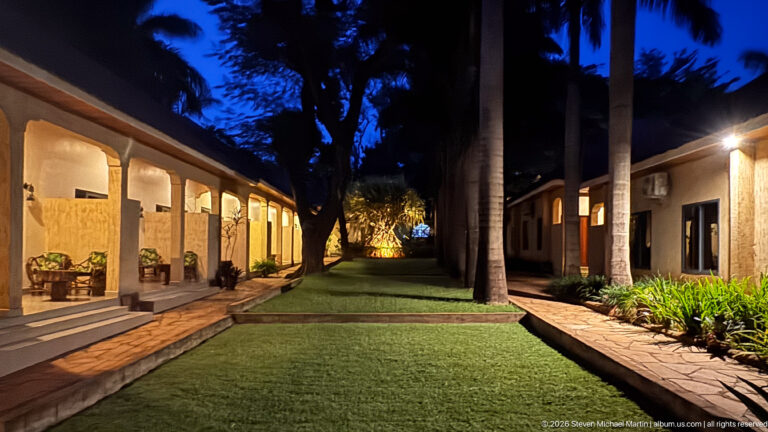 Guesthouse courtyard at night with lit verandas, rectangular lawn and tall palm trees under a deep blue sky.