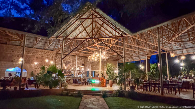Open-air restaurant pavilion at night with timber roof trusses, hanging lights, tables and surrounding plants.