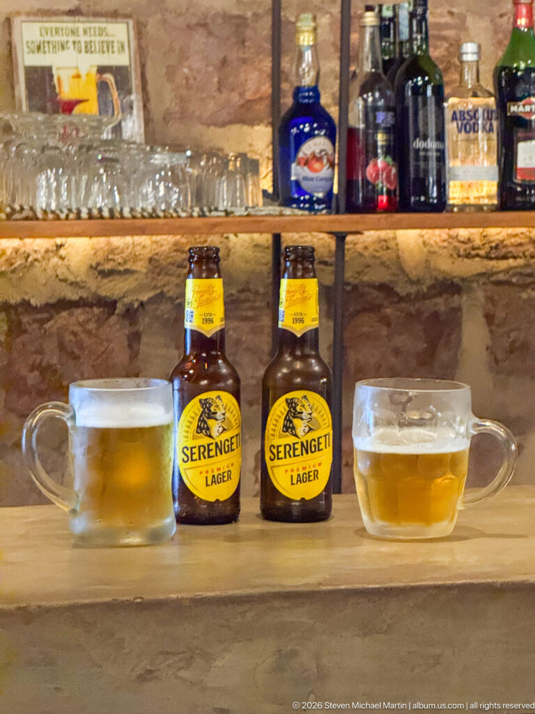 Two Serengeti Lager bottles and two handled glasses of golden beer on a rustic bar counter.