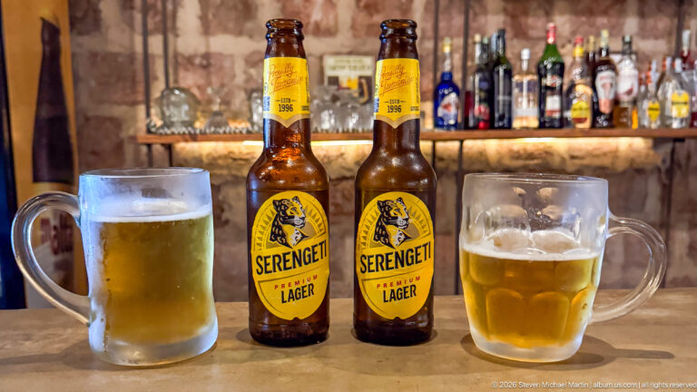 Two Serengeti Premium Lager bottles between frosted mugs of pale beer on a bar counter.