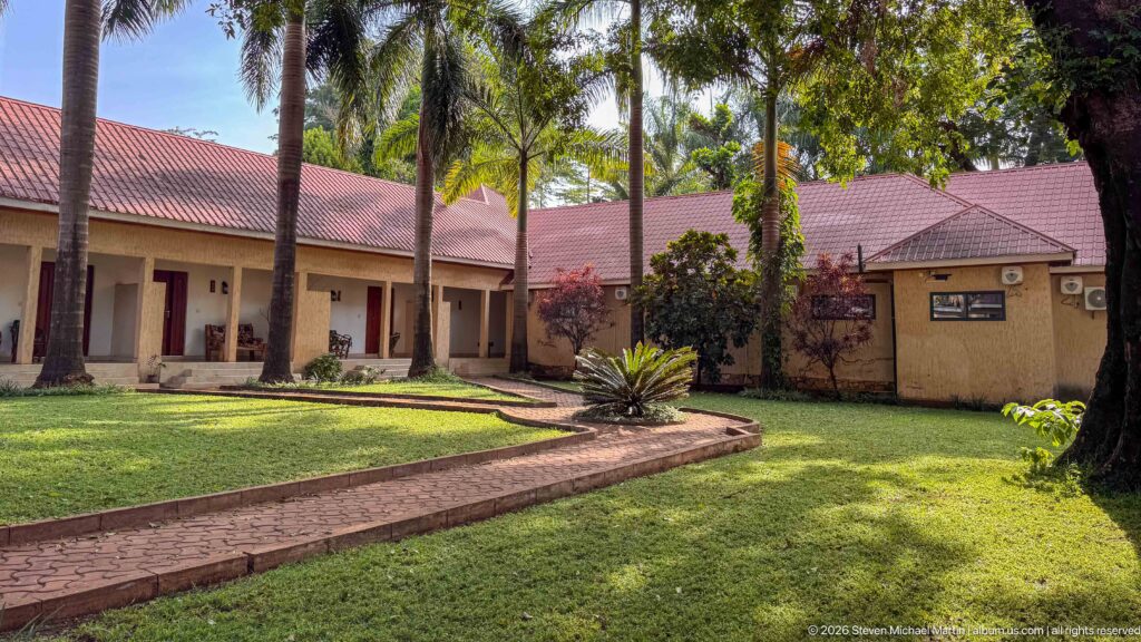 Cream guesthouse buildings with red roofs surround a green courtyard shaded by palms and mature trees.