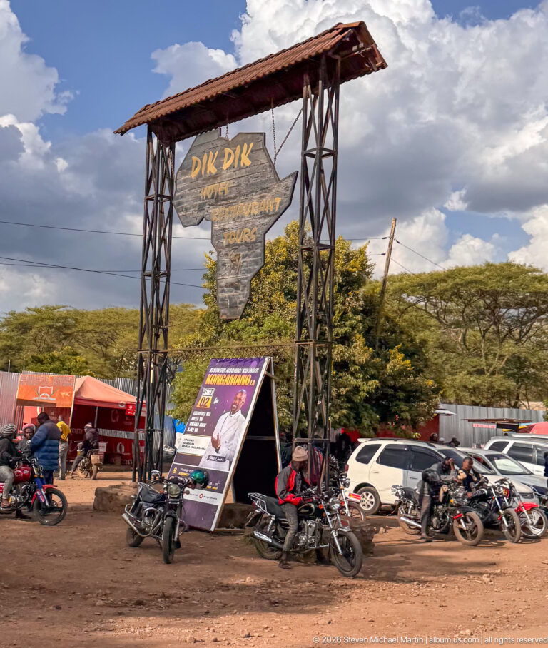 Large Dik Dik Hotel and Tours roadside sign above motorcycles, cars and promotional boards.