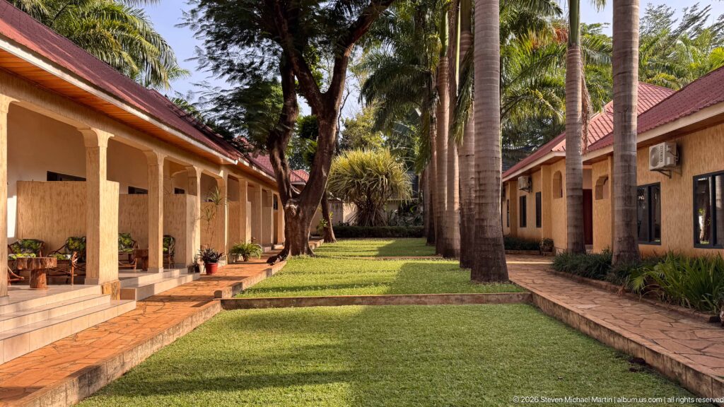 Long grassy courtyard between guesthouse wings, with a covered veranda, tall palms and paved walkways.