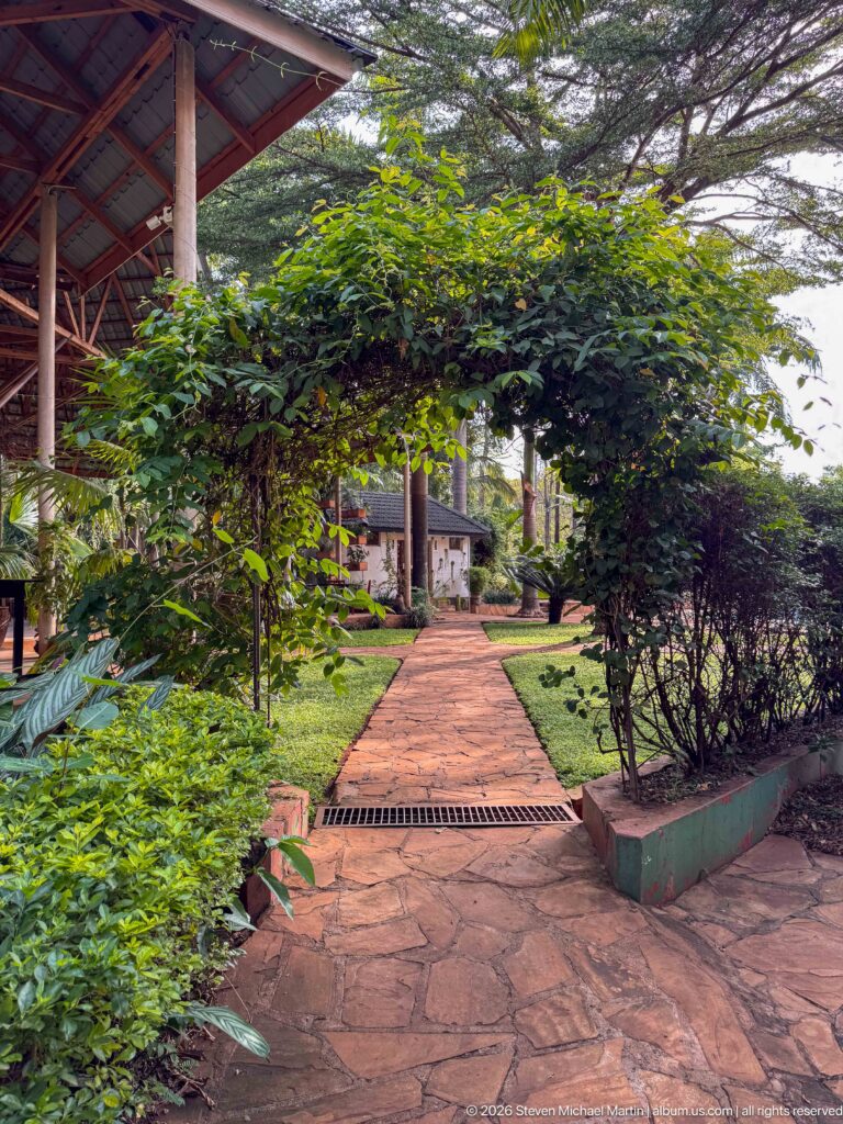Reddish paved walkway running through dense green lodge gardens with hedges, shrubs, trees, and roofed structures beyond.