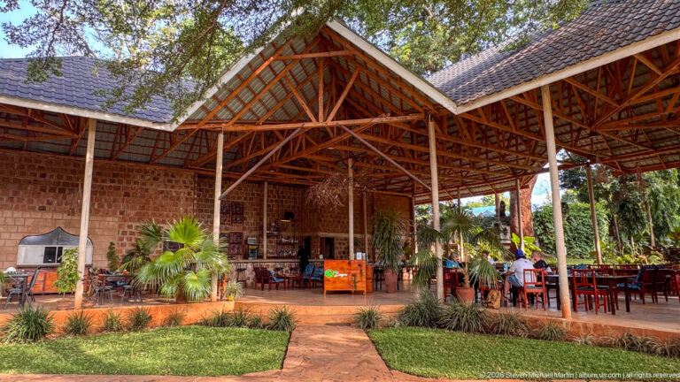 Open-sided brick pavilion with exposed timber roof trusses, dining tables, palms, lawn, and tiled roof sections.
