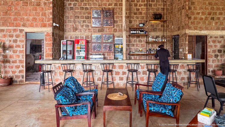 Open-air brick bar with high stools, beverage refrigerators, bottle shelves, and blue patterned lounge chairs around a low table.
