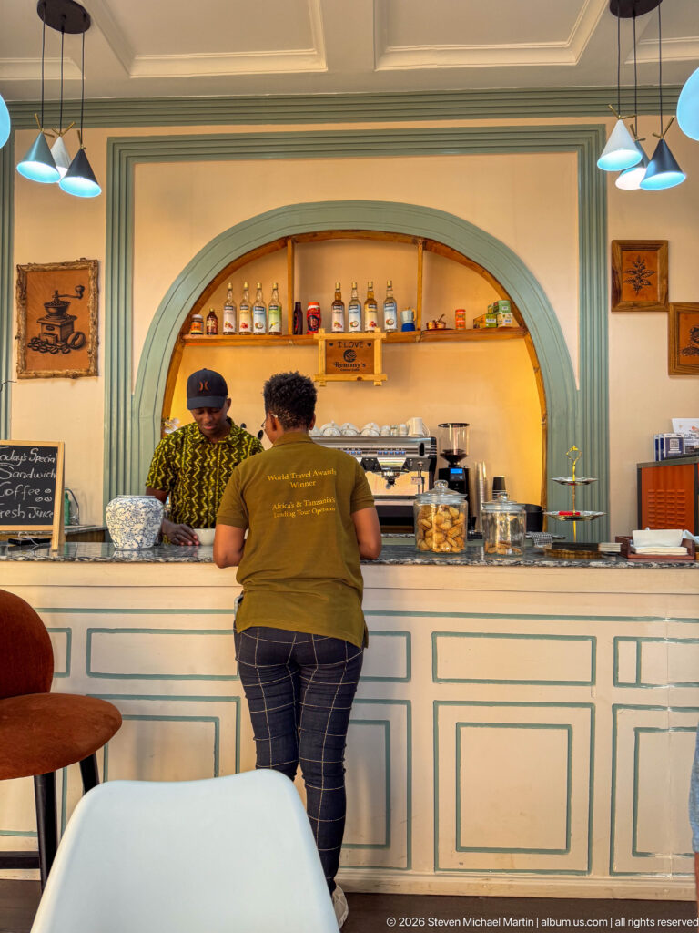 Two people at a pale cafe counter beneath arched shelves holding bottles and jars, with pendant lights overhead.