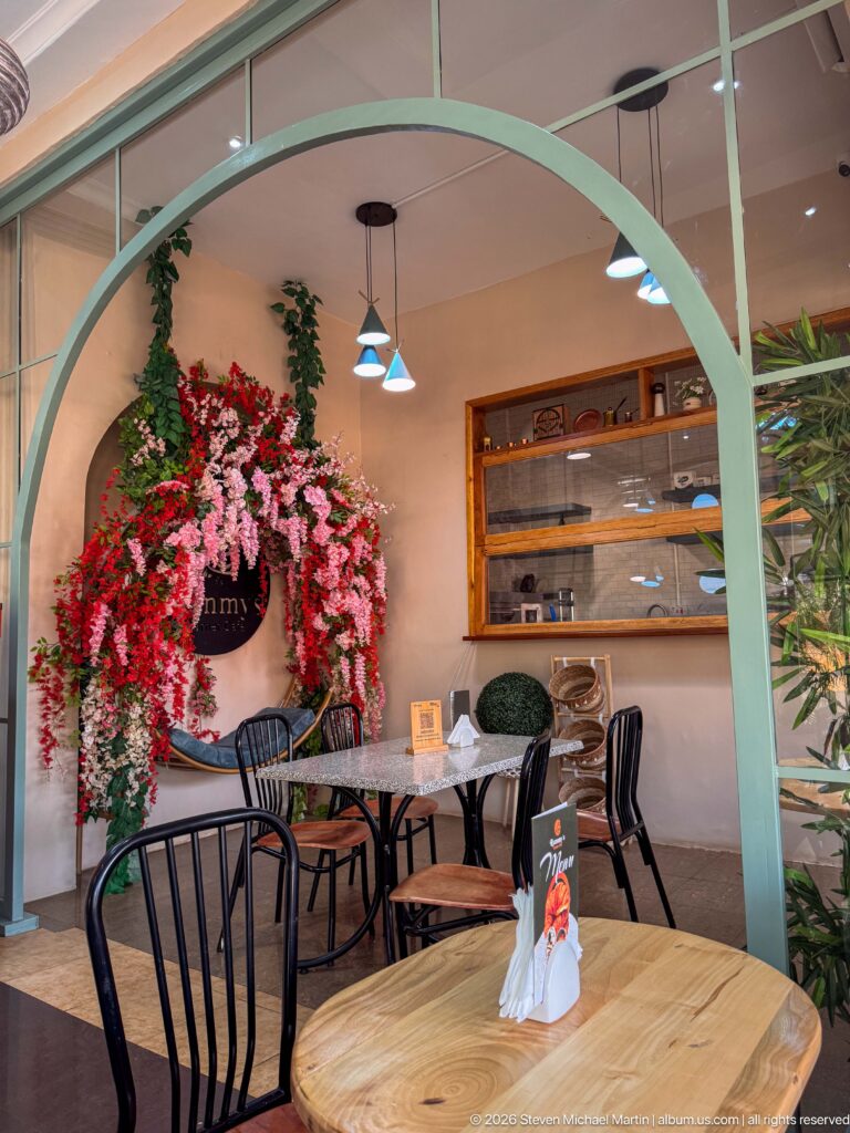 Cafe dining room with pale-green arches, wooden tables, black chairs, pendant lights, and cascading pink and red flowers.