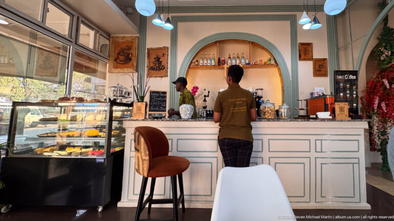 Customer at a pale cafe counter beside a pastry case, with staff, arched shelving, pendant lights, and floral decor.