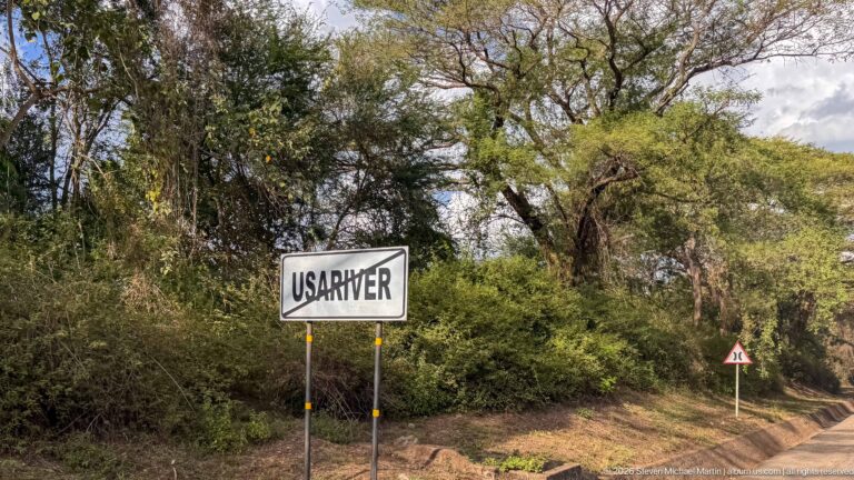 Roadside sign reading USA RIVER beside dense trees and shrubs, with a traffic warning sign farther along the road.