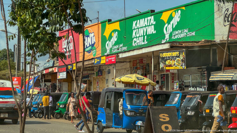 Moshi commercial street with colorful shop signs, pedestrians, and blue and green auto rickshaws parked along the curb.