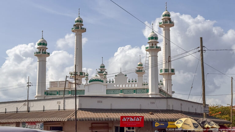 White mosque with four tall green-accented minarets rising above shops and utility wires in Moshi.