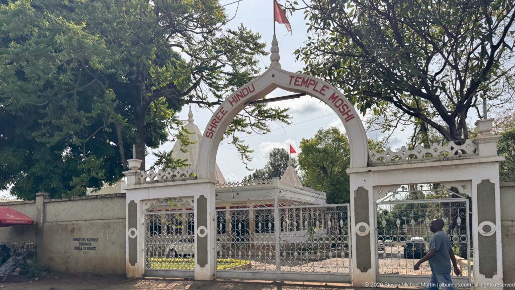 White arched entrance gate labeled Shree Hindu Temple Moshi, with metal gates, finials, trees, and the compound beyond.