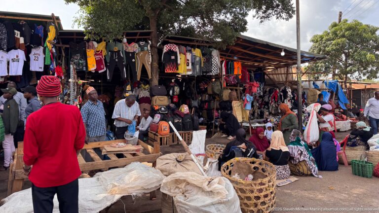 Busy open-air market with hanging clothes, baskets, bundled goods, seated vendors, shoppers, and a man in a red shirt.