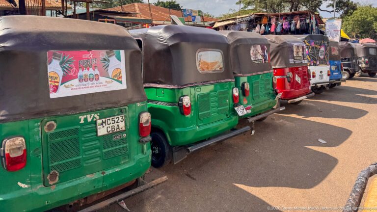 Row of green, red, blue, and black auto rickshaws parked closely along a sunlit market street.