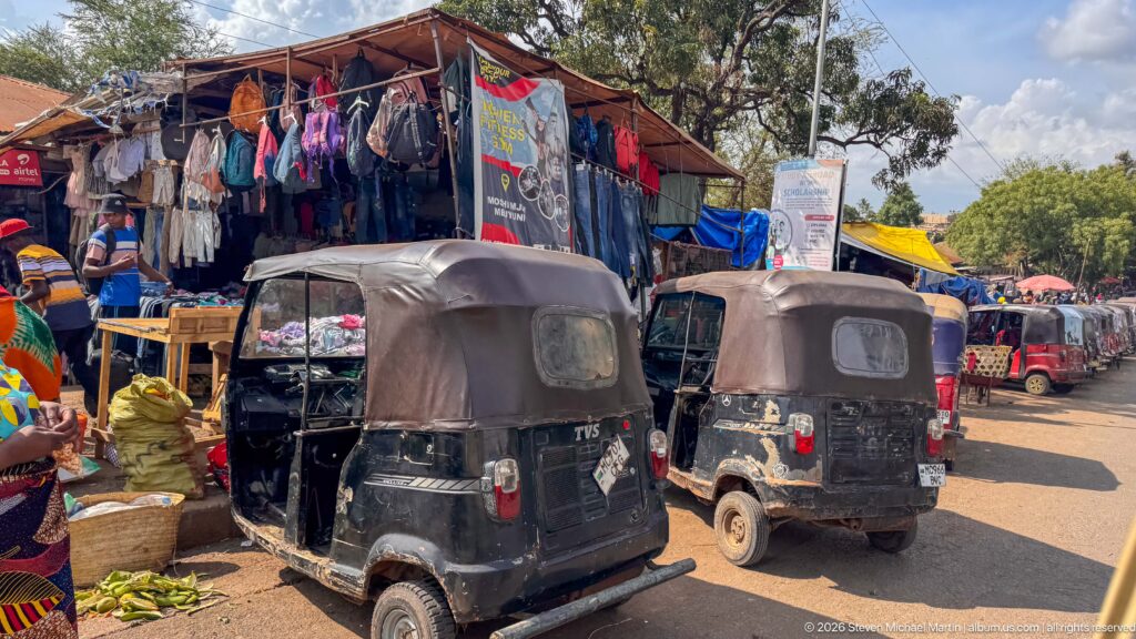 Two black auto rickshaws beside a clothing stall, baskets, and produce, with more rickshaws along the road.
