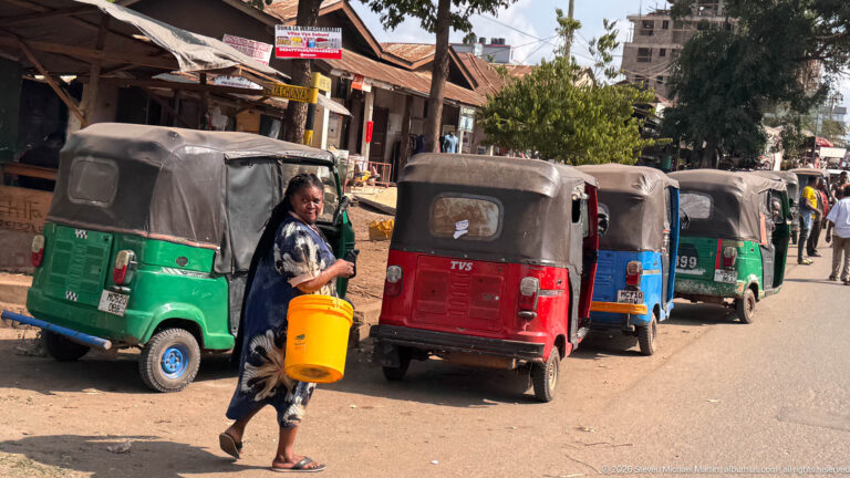 Woman carrying a yellow bucket beside a row of green, red, and blue auto rickshaws on a commercial street.