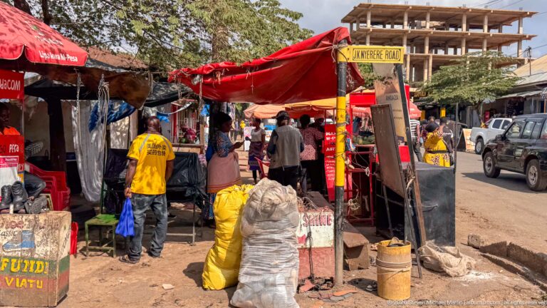 Roadside market stalls under red tarpaulins beside a Kitembe Road sign, with vendors, sacks, and containers.