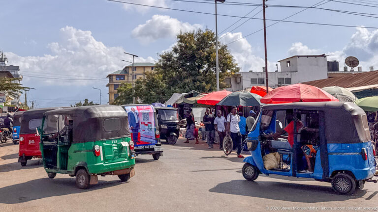 Green and blue auto rickshaws driving along a busy street with pedestrians, stalls, and umbrellas.