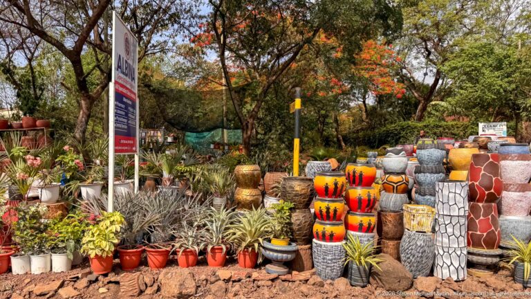 Roadside nursery with rows of potted plants and tall brightly painted decorative planters beneath trees.
