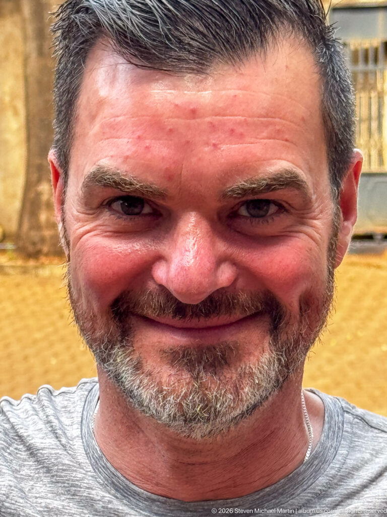 Close-up frontal portrait of a smiling bearded man in bright daylight.