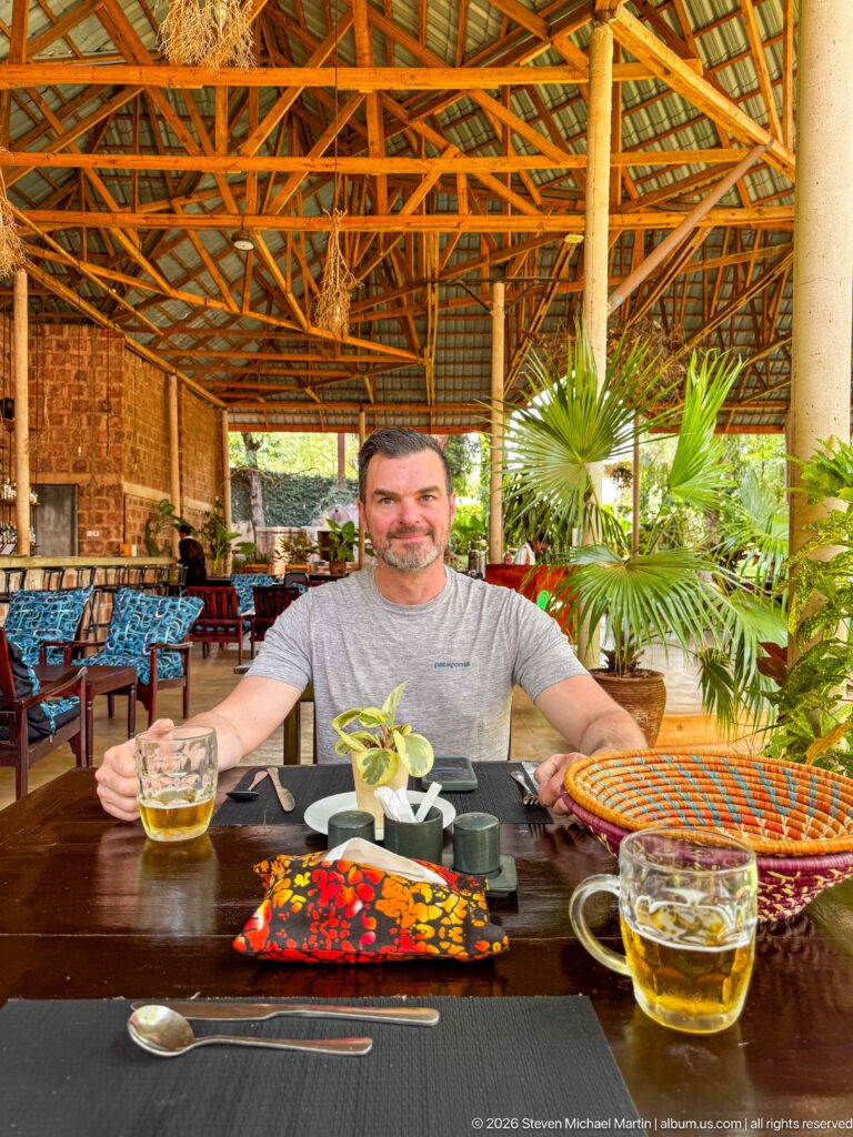 Man seated at a restaurant table with two amber drinks, condiments, flower, cutlery, and colorful woven accessories.