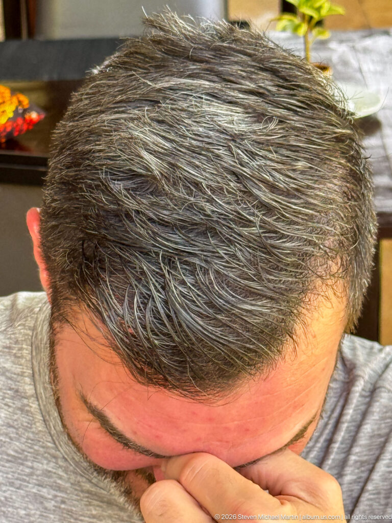 Overhead close-up of a seated man with salt-and-pepper hair, head bowed and one hand raised across his face.
