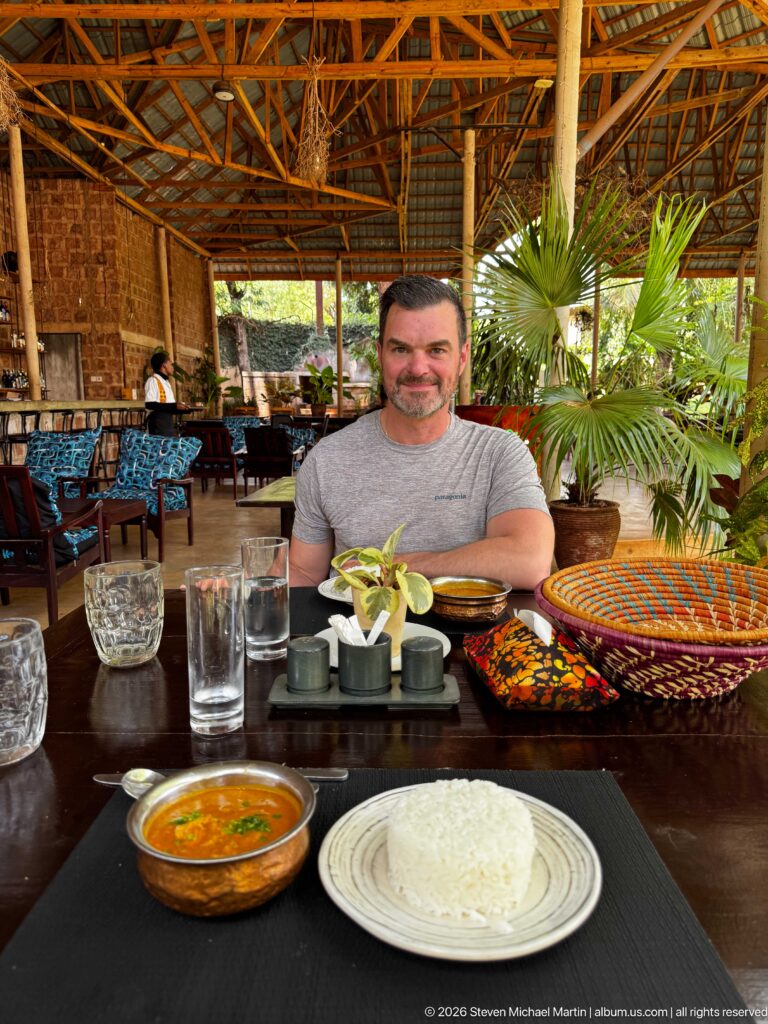 Man seated at an open-air restaurant table with a bowl of orange curry, white rice, glasses, and woven tableware.