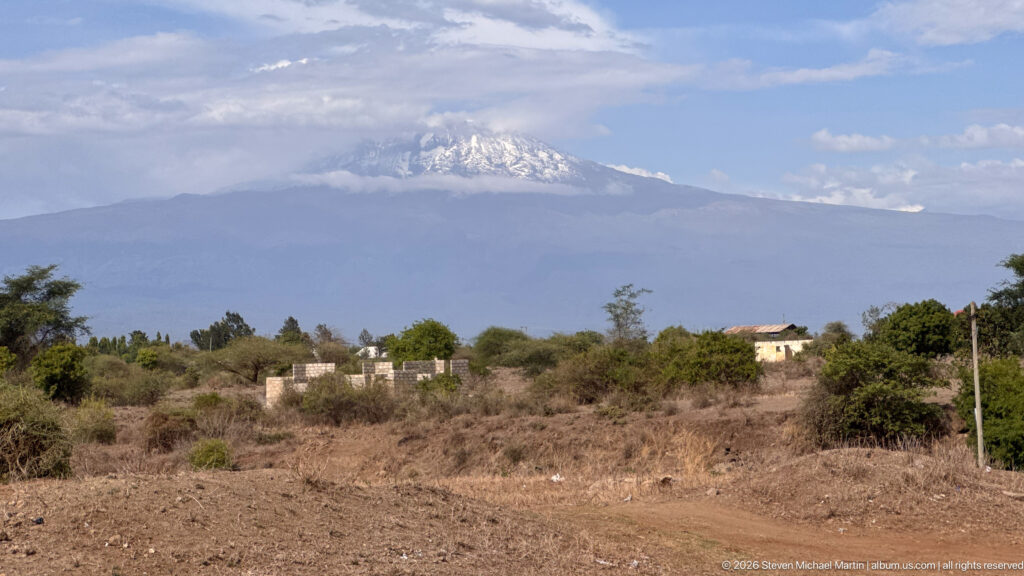 Distant snow-marked Mount Kilimanjaro above dry scrubland, scattered trees, rough ground, and a few low buildings.