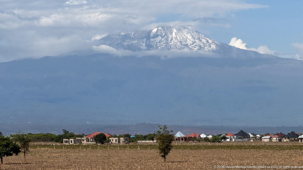 Snow-streaked summit of Mount Kilimanjaro rising through clouds above dry fields, scattered trees, and low houses.