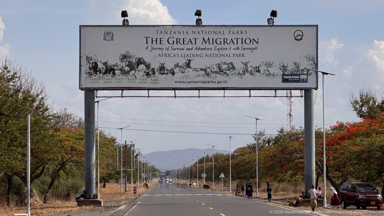 Large Tanzania National Parks Great Migration billboard above a broad tree-lined road with streetlights and distant hills.