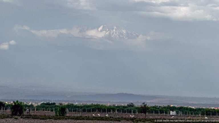 Hazy Mount Kilimanjaro above rows of cultivated vegetation, fencing and distant lowland settlement.