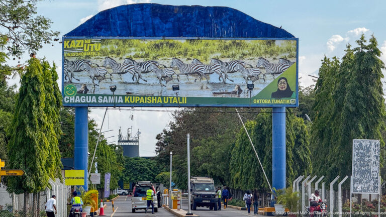 Roadway passing beneath a colorful gateway decorated with zebras and Swahili tourism text, with vehicles and pedestrians below.