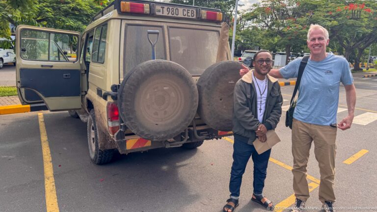 Two men standing beside the rear of a dusty safari vehicle with two spare tires in a paved parking area.