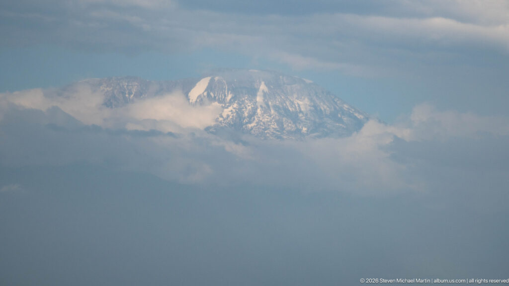 Distant snow-streaked summit of Mount Kilimanjaro emerging through layered blue-gray clouds.
