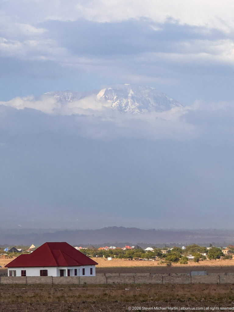 Cloud-wrapped Mount Kilimanjaro beyond open land, with a low white building and red roof in the foreground.