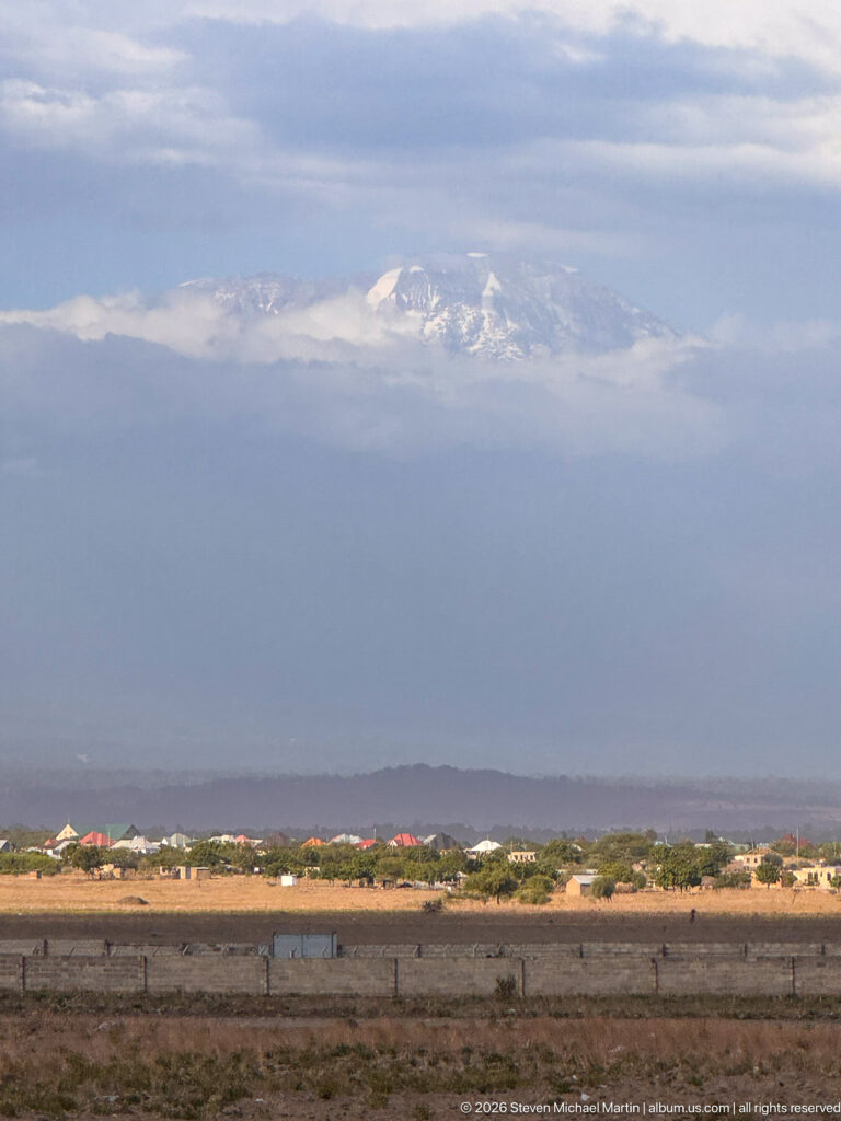 Mount Kilimanjaro beneath layered clouds beyond a dry plain and low settlement in the foreground.